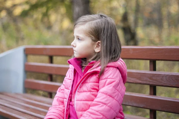 Kleines hübsches Mädchen im rosa Mantel, das im Herbst auf einer Holzbank im Park sitzt. Emotionales Porträt. Kindheitskonzept. Kaukasisch. Kind mit brünetten langen Haaren. Freizeit, Entspannung, Lebensstil — Stockfoto
