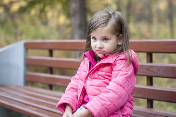 Kleines hübsches Mädchen im rosa Mantel, das im Herbst auf einer Holzbank im Park sitzt. Emotionales Porträt. Kindheitskonzept. Kaukasisch. Kind mit brünetten langen Haaren. Freizeit, Entspannung, Lebensstil — Stockfoto