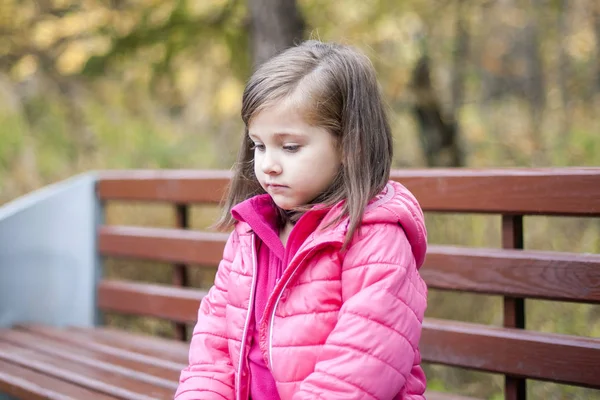 Маленькая симпатичная девочка в розовом пальто сидит на деревянной скамейке в парке осенью. Эмоциональный портрет. Концепция детства. Белый. Ребёнок с длинными волосами брюнетки. Отдых, релаксация, образ жизни — стоковое фото
