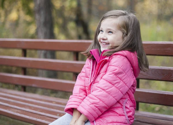 Маленькая симпатичная девочка в розовом пальто сидит на деревянной скамейке в парке осенью. Эмоциональный портрет. Концепция детства. Белый. Ребёнок с длинными волосами брюнетки. Отдых, релаксация, образ жизни — стоковое фото