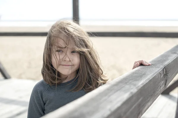 Vacation by the sea. Cute kid on the deserted beach. Summer, outdoors. Wind in the hair. Little smiling sweet baby girl. Beach landscape. On the blurred background of the wood railing and the sea