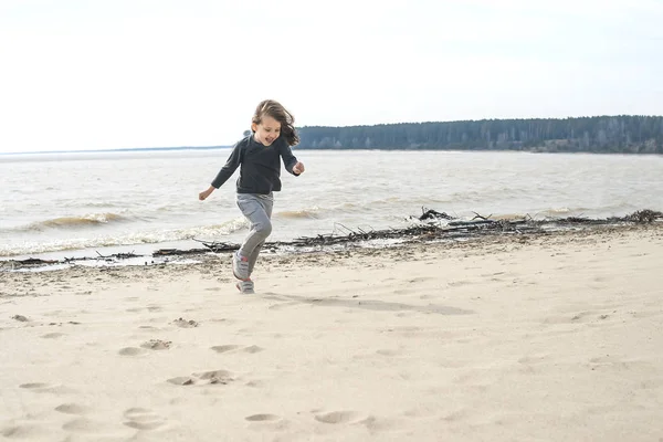 Pequena menina doce sorridente com cabelo castanho correndo e pulando na praia. Férias junto ao mar. Miúda gira na praia deserta. Verão, ao ar livre. Vento na rapariga do cabelo. Paisagem praia — Fotografia de Stock
