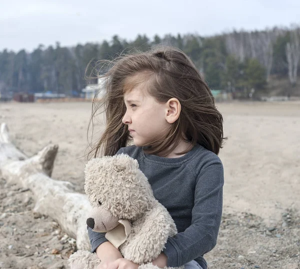 Lonely little girl feel stressed, hugs a teddy bear. Baby sitting on a old fallen tree on the beach and staring into the distance. Depression, helplessness, indifference, lostness, sadness. Windy day
