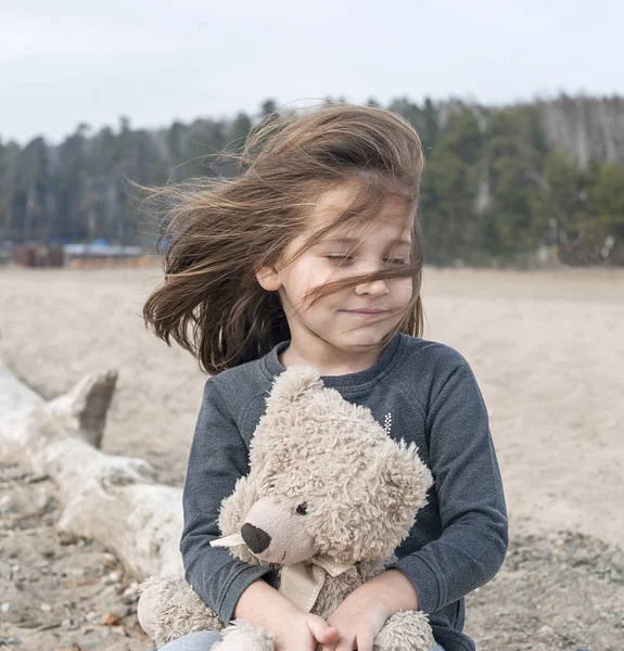 Mała dziewczynka siedzi na stare, Spasione drzewo, na plaży, przytula pluszowego misia. Zadowolenie, jazdy, entuzjazm, euforia, podmuch, radość, mimikry, pokoju, przyjemności, satysfakcji, serenity, tkliwość. Wietrzny dzień — Zdjęcie stockowe