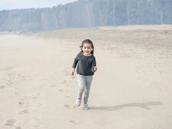Маленькая улыбающаяся милая девочка с каштановыми волосами бегает и прыгает по пляжу. Отпуск у моря. Симпатичная девочка на пустынном пляже. Саммер, на улице. Ветер в волосах девушки. Пляжный пейзаж — стоковое фото