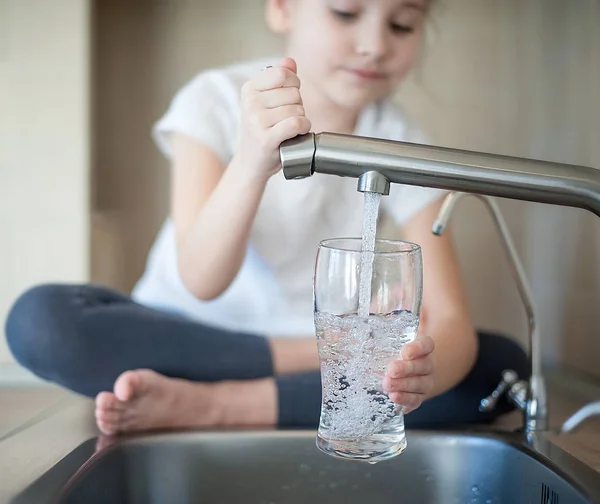 Kleines Mädchen öffnet einen Wasserhahn mit der Hand, die ein Glas hält — Stockfoto