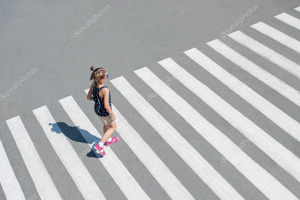 Stylish child in sun glasses, fashion clothes walking along summer city crosswalk. Kid on pedestrian side walk. Concept pedestrians passing a crosswalk. From top view. Behind. Shadow at zebra crossing