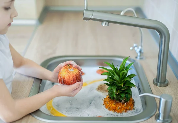 Child is washing an apple above kitchen sink. Washing and sanitizing fruits and vegetables in soapy water. Decontamination and disinfection of products from the store. Coronavirus infection prevention