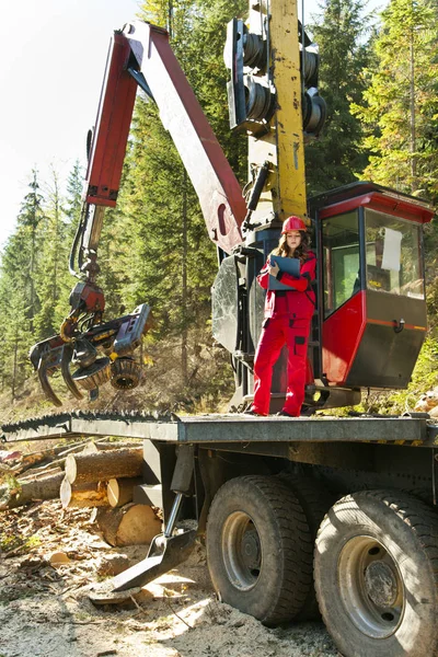 Young forest engineer standing in front of forestry machinery for logging and writing to notepad. She is wearing red work clothes and a helmet.