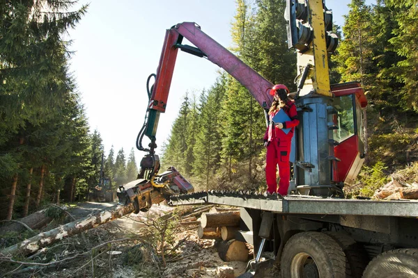 Young forest engineer standing in front of forestry machinery for logging and writing to notepad. She is wearing red work clothes and a helmet.