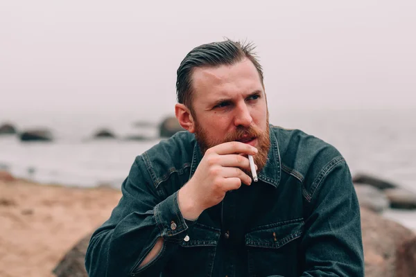 Grave uomo barbuto fumare una sigaretta sulla spiaggia — Foto Stock
