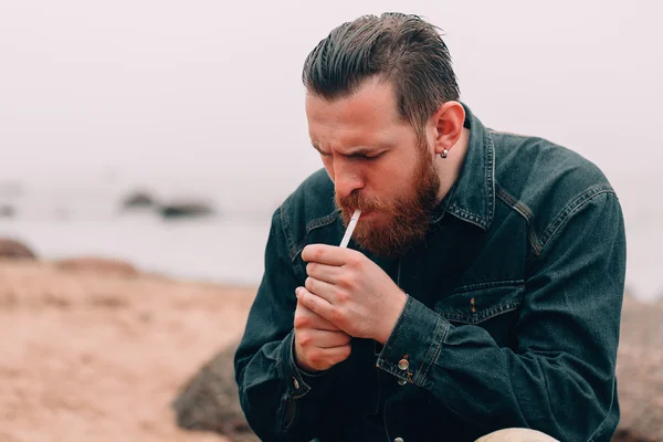 Серьезный бородатый мужчина курит сигарету на пляже — стоковое фото