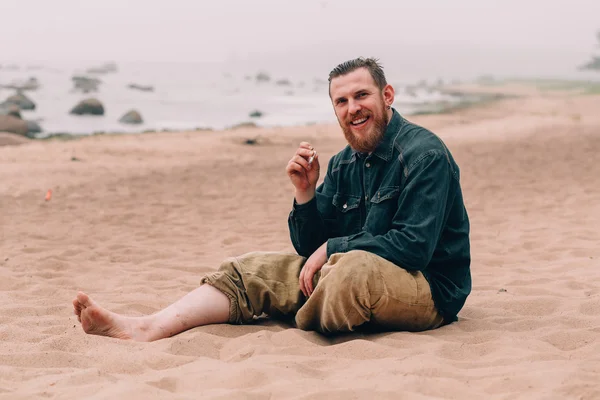 Grave uomo barbuto fumare una sigaretta sulla spiaggia — Foto Stock