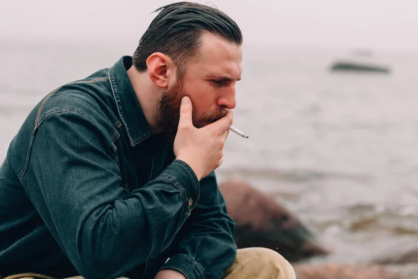Серьезный бородатый мужчина курит сигарету на пляже — стоковое фото