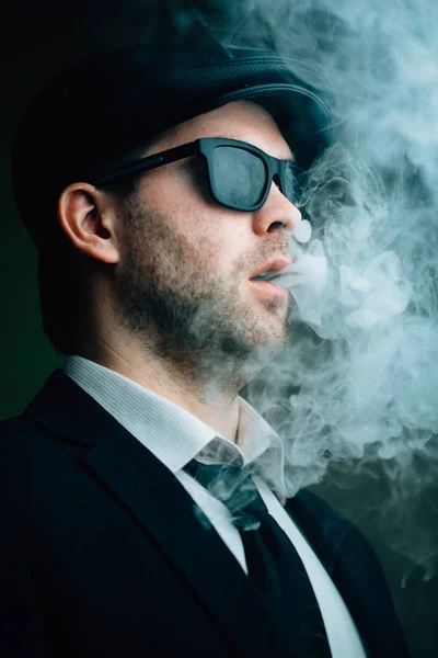 Модный мужчина в солнцезащитных очках и кожаной кепке выдыхает белый дым — стоковое фото