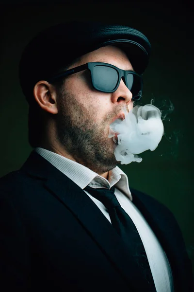 Módní člověk v sluneční brýle a koženou čepici vydechuje bílý kouř — Stock fotografie