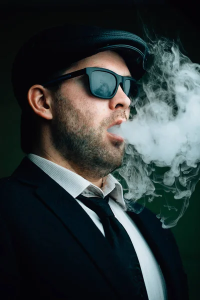 Модный мужчина в солнцезащитных очках и кожаной кепке выдыхает белый дым — стоковое фото