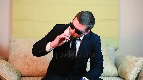 Stijlvolle jonge zakenman rookt een elektronische sigaret op een sofa — Stockvideo