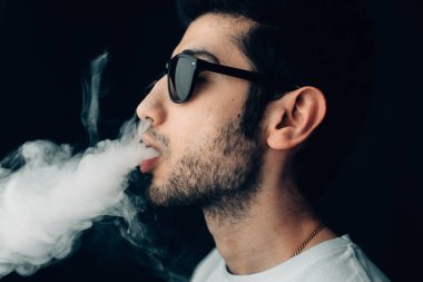 Güneş gözlüklü genç cool adam bir duman bulutu exhales. Profil yakın çekim yatay portre Studio.