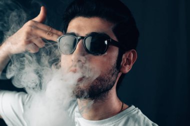Güneş gözlüklü genç cool adam onun tapınağa parmak ile duman bulutu exhales. Stüdyo yakın çekim yatay dikey.