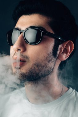 Güneş gözlüklü genç cool adam bir duman bulutu exhales. Dikey stüdyo yakın çekim portre.