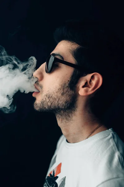 Young cool guy in sunglasses exhales a cloud of smoke. Vertical studio portrait in profile close-up. — Stock Photo, Image