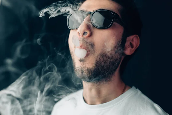 Jovem cara legal em óculos de sol exala uma nuvem de fumaça. Estúdio retrato horizontal em close-up . — Fotografia de Stock