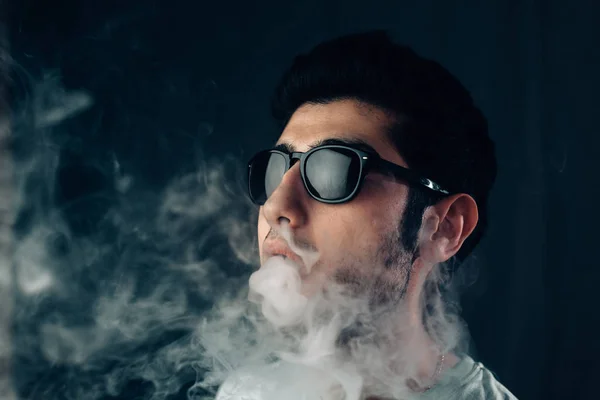 Jeune gars cool dans des lunettes de soleil exhale un nuage de fumée. Portrait horizontal studio en gros plan . — Photo