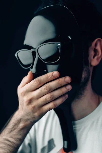 Homem de máscara preta brilhante e óculos de sol fecha a boca com a mão. Retrato de estúdio vertical em close-up . — Fotografia de Stock