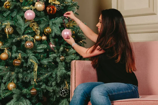 Hermosa joven morena en un sofá decora el árbol de Navidad. Estudio horizontal retrato . —  Fotos de Stock