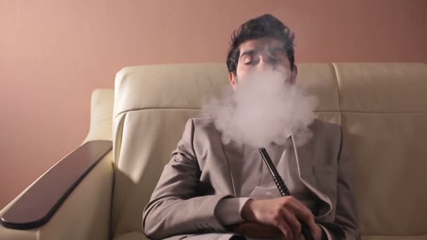 Empresário fumando um narguilé e relaxando em um sofá no quarto acolhedor . — Vídeo de Stock