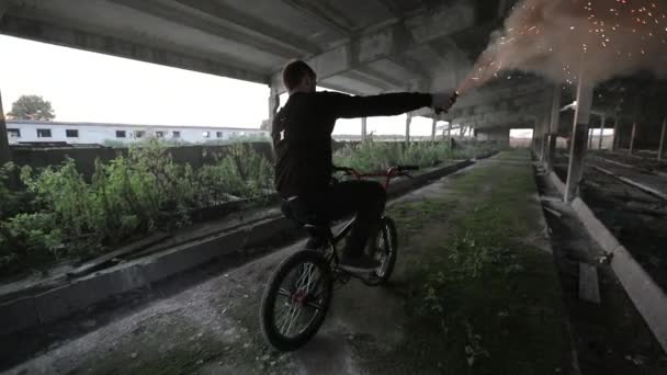 Велосипедист с оранжевым дымом в руке проезжает по заброшенному зданию — стоковое видео