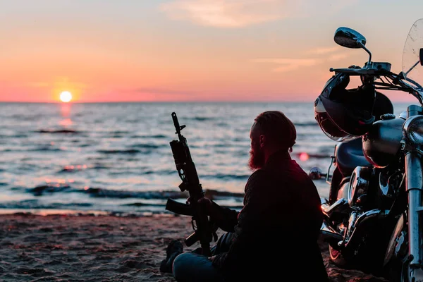 Biker mit automatischen Waffen in der Nähe eines Fahrrads am Strand bei Sonnenuntergang — Stockfoto