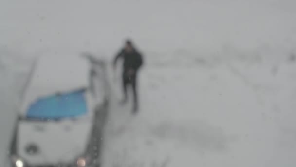 Schnee fällt auf dem Hintergrund eines Mannes, der sein Auto auf dem Parkplatz umgräbt — Stockvideo