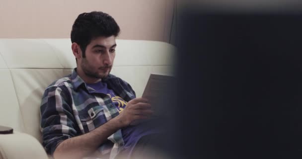 Junger Mann liest aufmerksam ein Buch, das auf der Couch liegt — Stockvideo