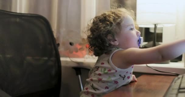 Маленькая девочка с вьющимися волосами и пустышкой во рту, сидящей рядом с монитором компьютера — стоковое видео