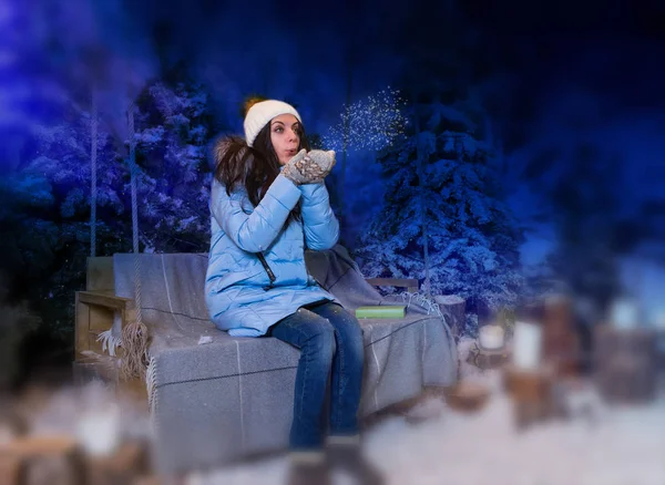 Frau in blauer Daunenjacke pustet Schneeflocken, während sie auf einem sw sitzt — Stockfoto