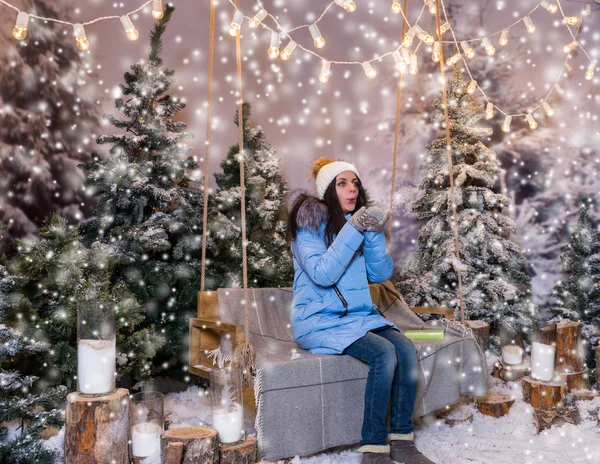 Kvinnan i blå dunjacka blåser snöflingor medan du sitter på en sw — Stockfoto