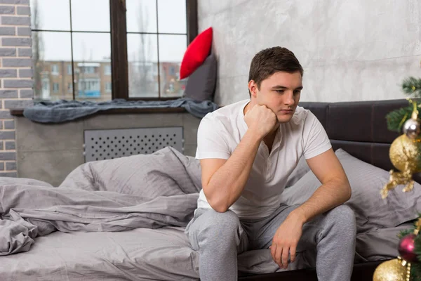 Young man is leaning on his hand while sitting sadly on bed, wom — Stock Photo, Image