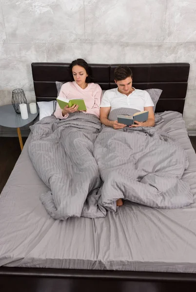 Young concentrated pair reading books while lying in the bed and — Stock Photo, Image
