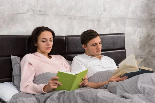 Молодая счастливая пара читает книги, лежа в постели и нося — стоковое фото