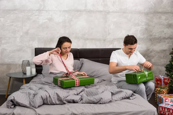 Pareja joven en pijama desempacando sus regalos mientras están sentados o — Foto de Stock