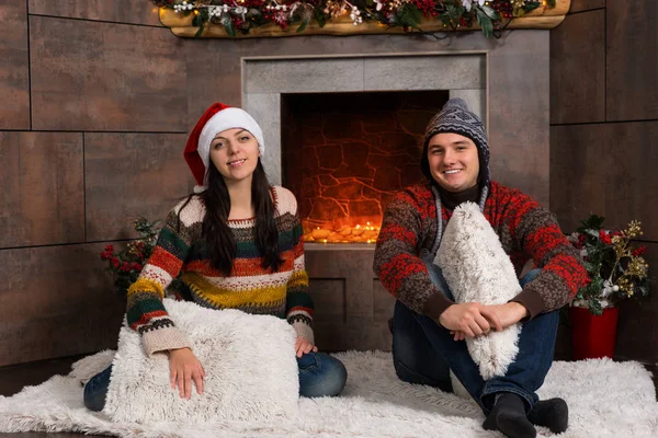 Joven pareja sonriente en divertidos sombreros de invierno sentado en una alfombra en fr —  Fotos de Stock