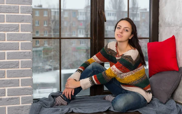 Smiling young woman in sweater sitting on the windowsill and loo — Stock Photo, Image