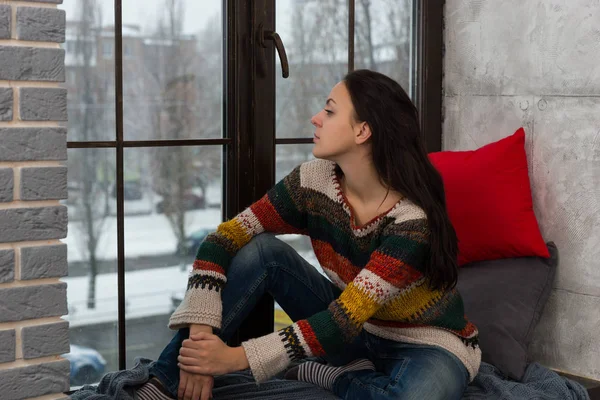 Thoughtful young woman in sweater sitting on the windowsill and — Stock Photo, Image