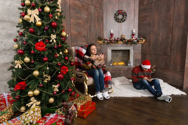 Pareja joven sentada en la habitación con árbol de Navidad y sirviendo —  Fotos de Stock