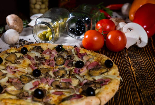 Pizza con prosciutto e olive nere sul tavolo di legno e altre verdure — Foto Stock