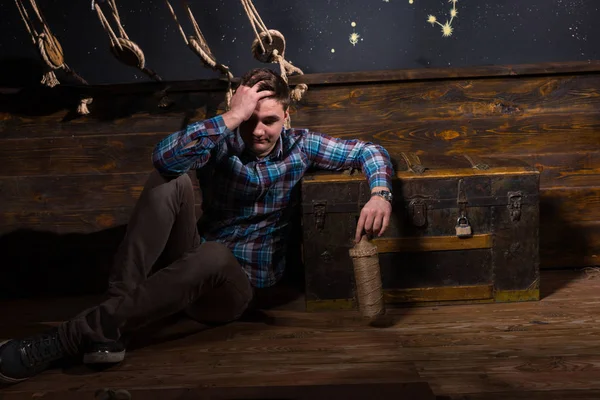 A young disappointed man sits near a chest, holding glass bottle — Stock Photo, Image