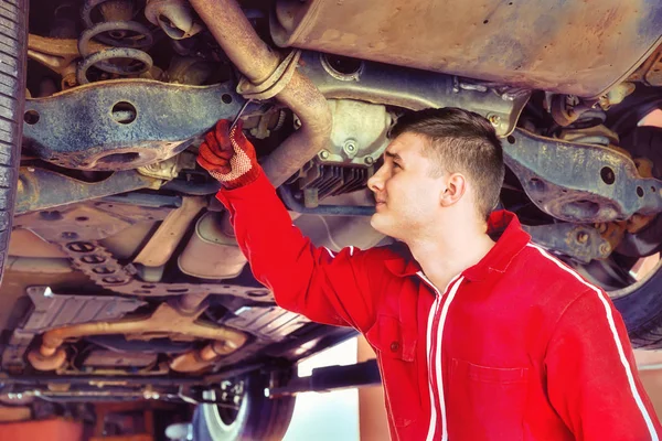 Jovem mecânico de automóveis em uniforme trabalhando sob um carro levantado w — Fotografia de Stock
