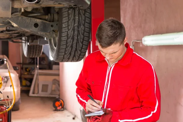 Young male auto mechanic standing making notes underneath a lift
